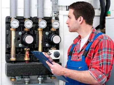 HVAC technician in a plaid shirt with blue coveralls standing with a clipboard, inspecting a boiler.