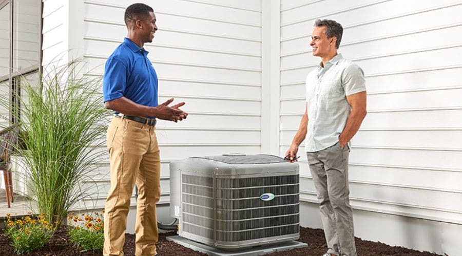 HVAC technician and a homeowner stand beside an air conditioning unit beside a white house